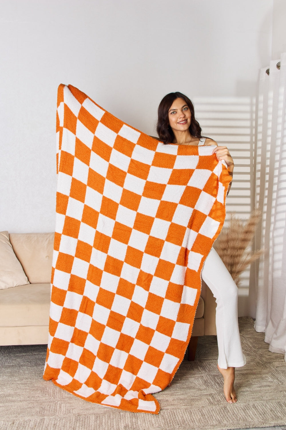 A person stands holding a large Cuddley Checkered Decorative Throw Blanket, smiling, in a room with a sofa and a potted plant. The blanket features a black and white checkerboard pattern, is made from 100% polyester, and imported for quality.