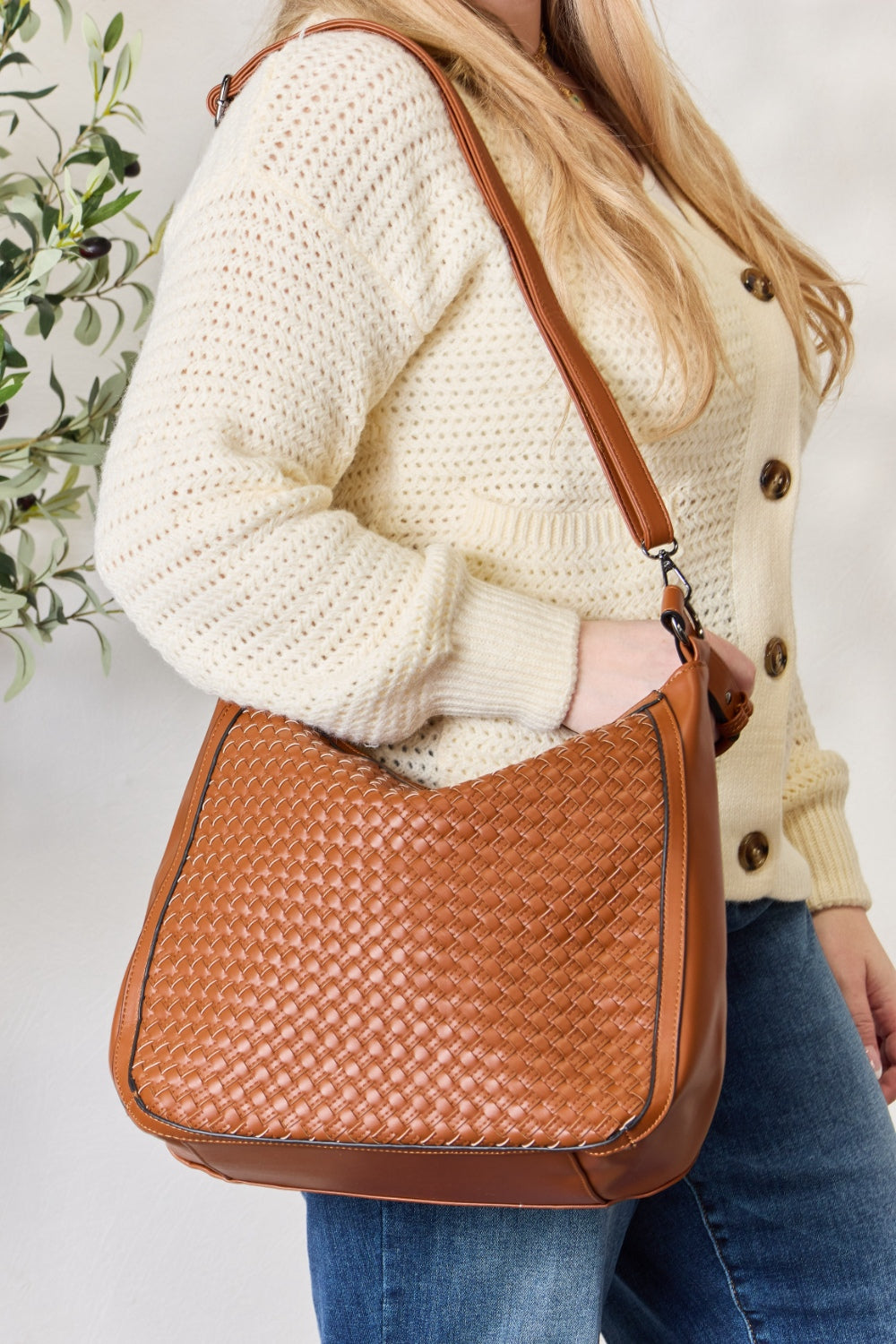 A person carries the SHOMICO Weaved Vegan Leather Handbag in a stylish brown hue, featuring a sleek crossbody strap, expertly paired with a cream sweater and jeans.