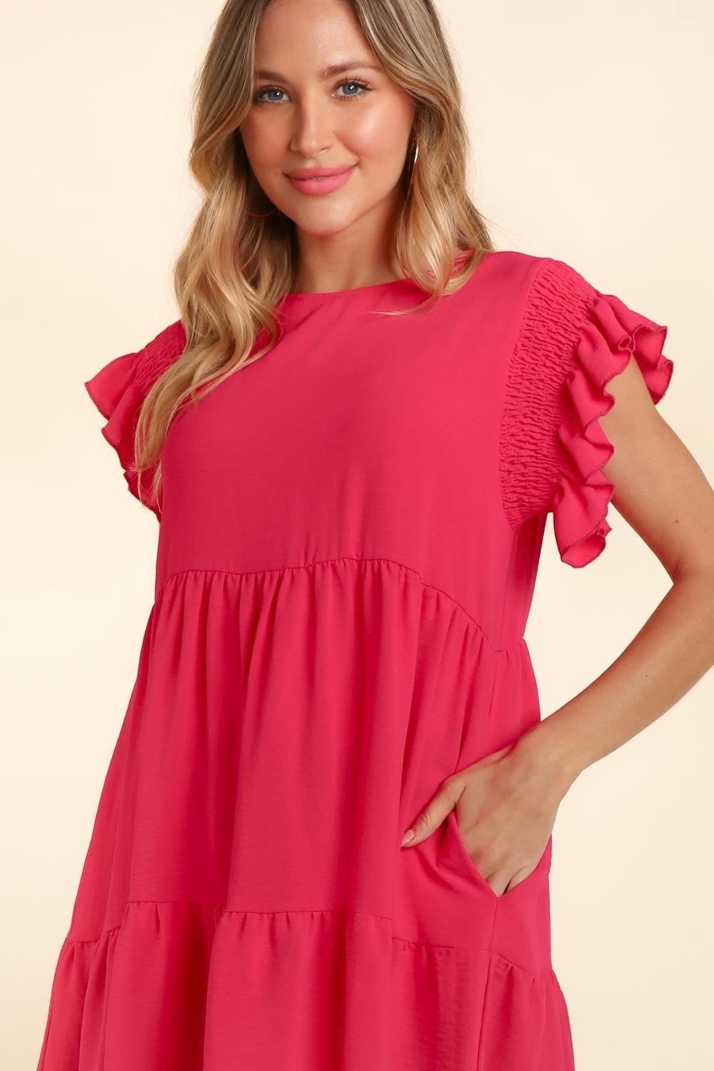 A woman with long, wavy hair wearing a bright pink Haptics Full Size Smocking Ruffle Short Sleeve Dress with Pockets, featuring ruffled sleeves and a smocked waist, stands smiling with arms crossed in front of a neutral background.