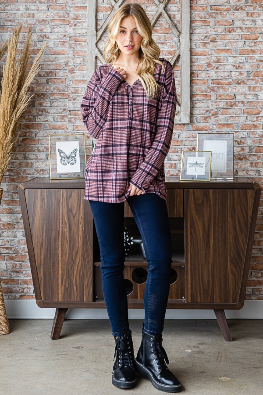 Sporting long blonde hair, a person stands against a brick wall wearing the Heimish Full Size Plaid V-Neck Long Sleeve Top in pink and black. Framed butterfly prints adorn the background, while this adaptable wardrobe item adds a touch of flair to any ensemble.