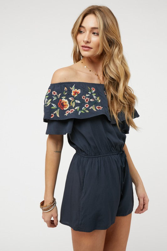 Wearing the stylish Floral Embroidered Off Shoulder Romper, a woman poses gracefully, showcasing its ruffle detail and making it an ideal addition to any summer wardrobe.