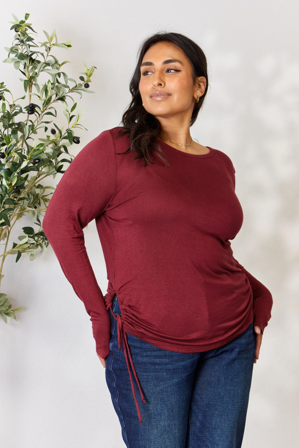 A woman stands in front of a plain background and a small tree, wearing the Culture Code Full Size Drawstring Round Neck Long Sleeve Top in maroon and blue jeans.