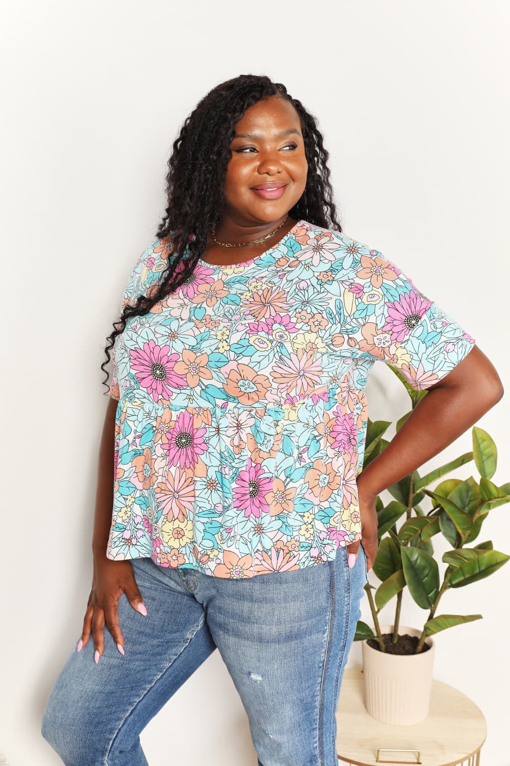 A person wearing a Double Take Floral Round Neck Babydoll Top and jeans stands with one hand in their pocket, with a plant in the background.