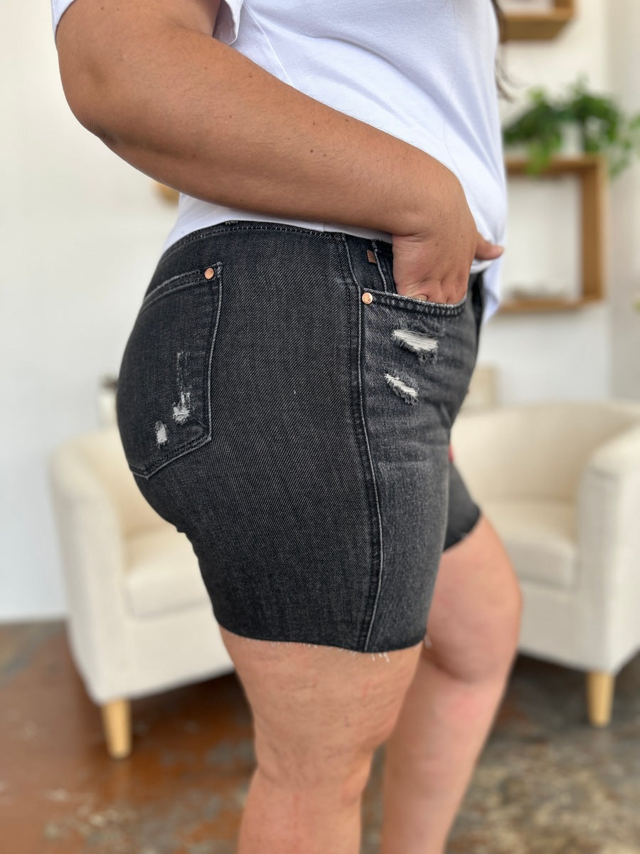 A person wearing a white t-shirt and Judy Blue Full Size High Waist Rigid Magic Denim Shorts is standing indoors in front of a beige chair.