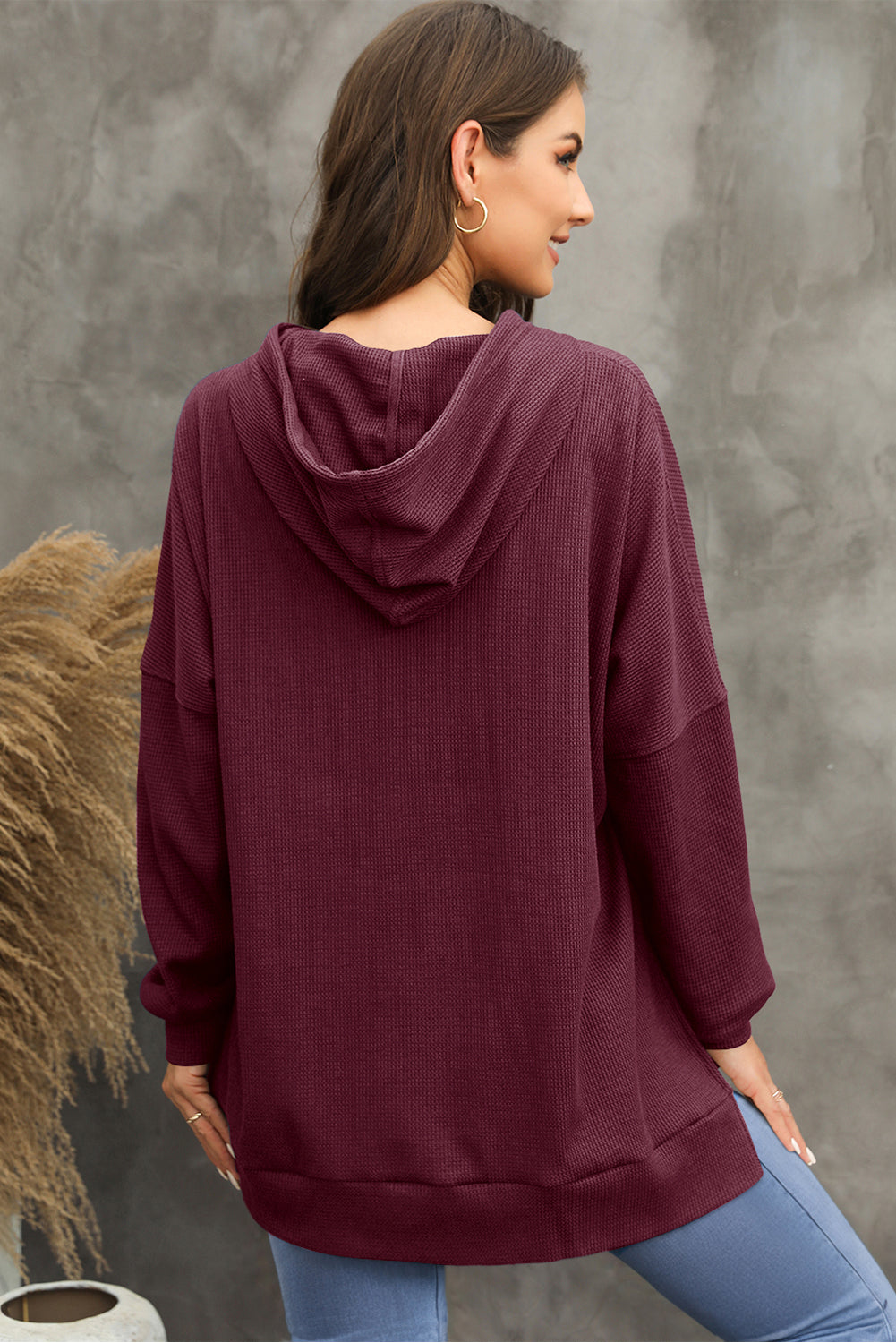 A woman in a Red Dahlia Waffle Knit Fleece Lined High Low Oversized Hoodie and black shorts is seen from behind, standing against a plain gray background.