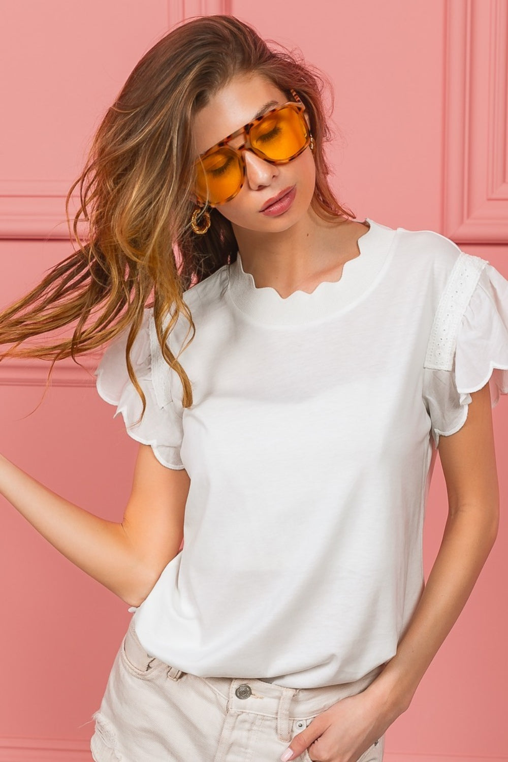 A woman with wavy hair dons the elegant BiBi Scallop Edged Lace Trim Top and large amber sunglasses while standing in front of a vibrant pink wall.