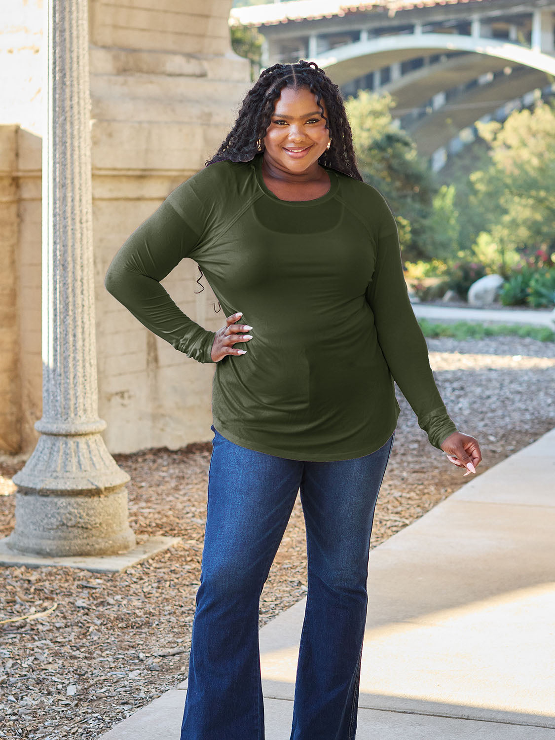 A woman wearing sunglasses and a slightly stretchy Basic Bae Full Size Round Neck Long Sleeve T-Shirt in white stands on a sidewalk with an arched structure in the background, smiling slightly.