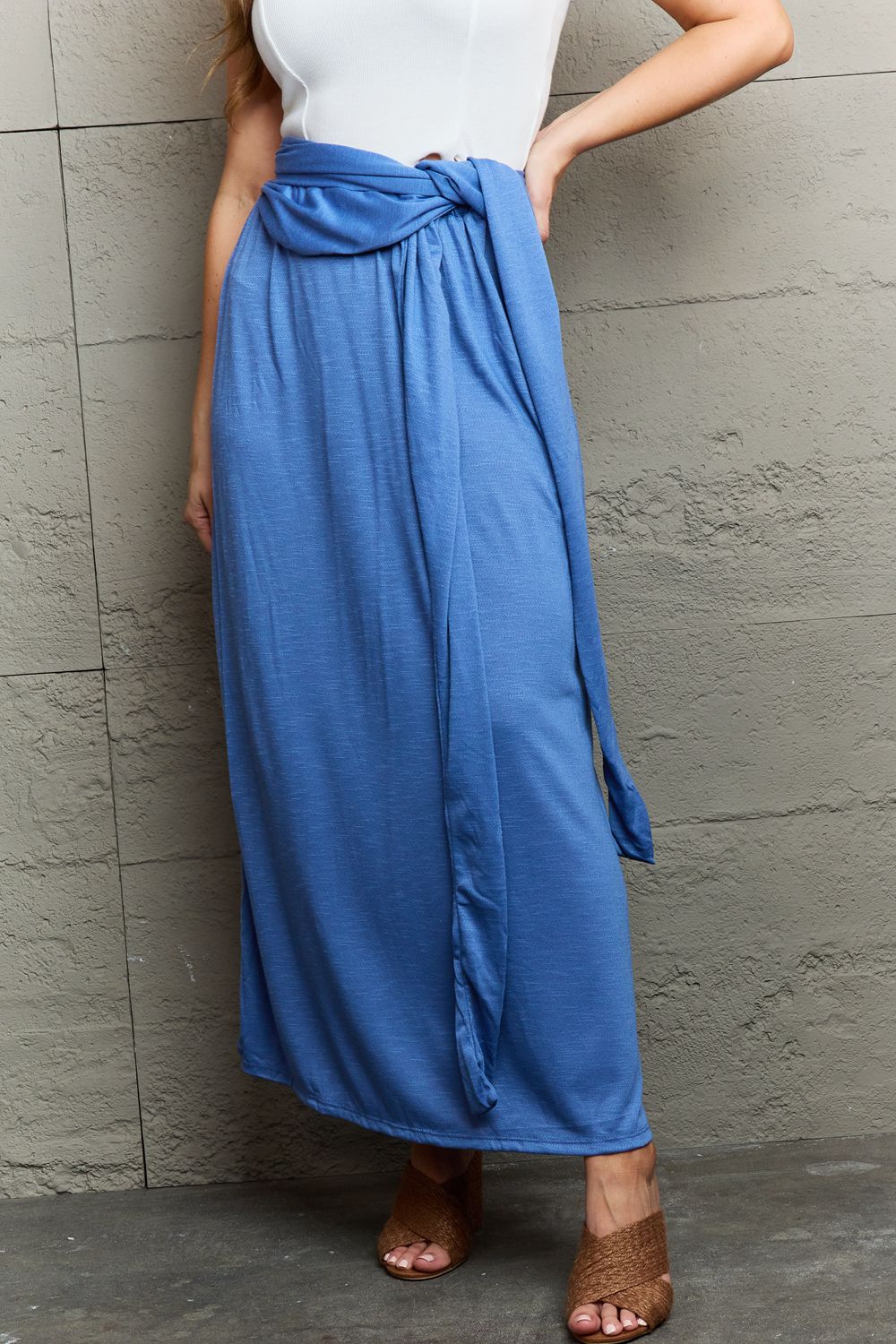 A person stands against a textured gray wall, smiling in the Ninexis Know Your Worth Criss Cross Halter Neck Maxi Dress in blue, featuring a stylish cut-out design. One hand rests on the wall while the other is confidently placed on their hip.