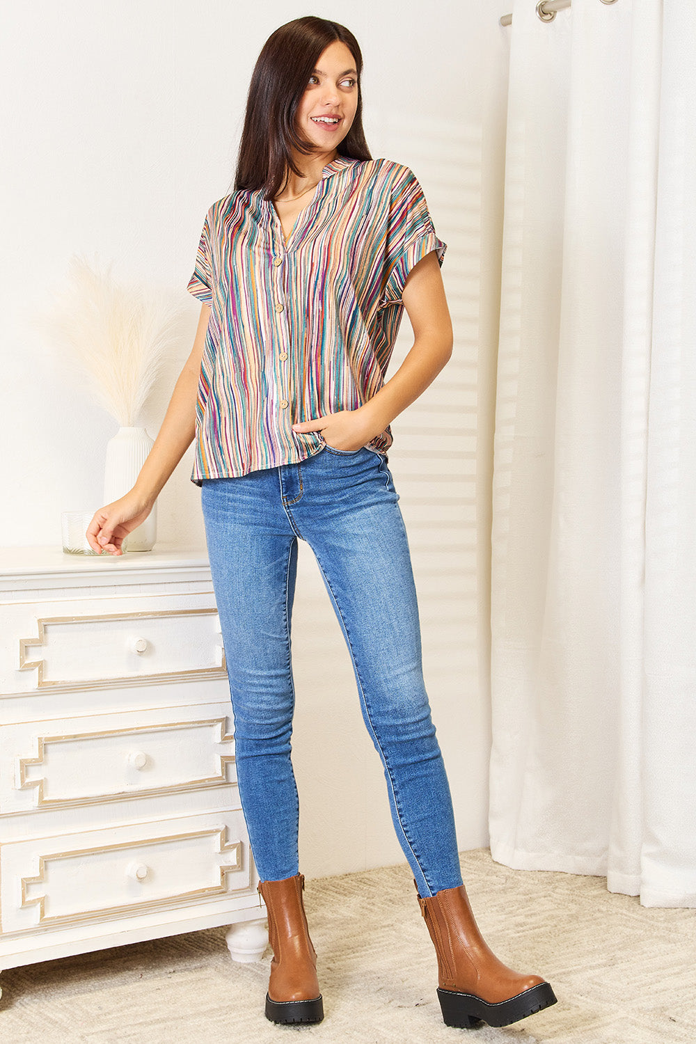 A person with long, curly hair wearing a Shiny Multicolored Stripe Notched Neck Top and blue jeans stands with one hand in their pocket, smiling slightly.