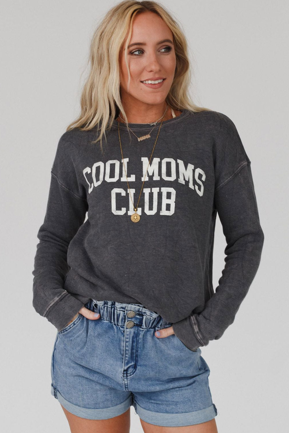 A person with long blond hair wearing a gray COOL MOMS CLUB drop shoulder sweatshirt and relaxed-fit blue jeans is seen from the back, standing near a pumpkin and a window.