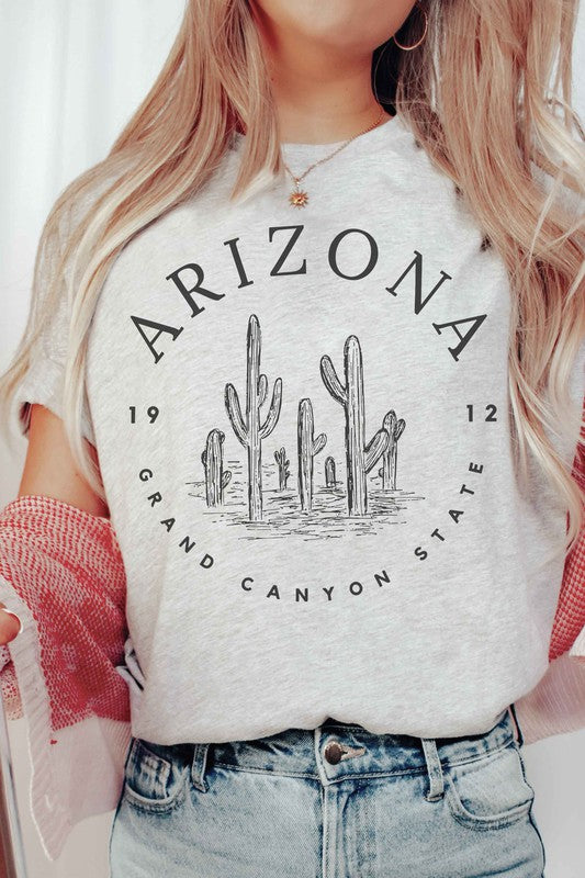 Person wearing an ARIZONA GRAND CANYON STATE Graphic Tee, made of premium 100% cotton with a design featuring the words "ARIZONA GRAND CANYON STATE 1912" and a drawing of cacti, paired with white shorts.