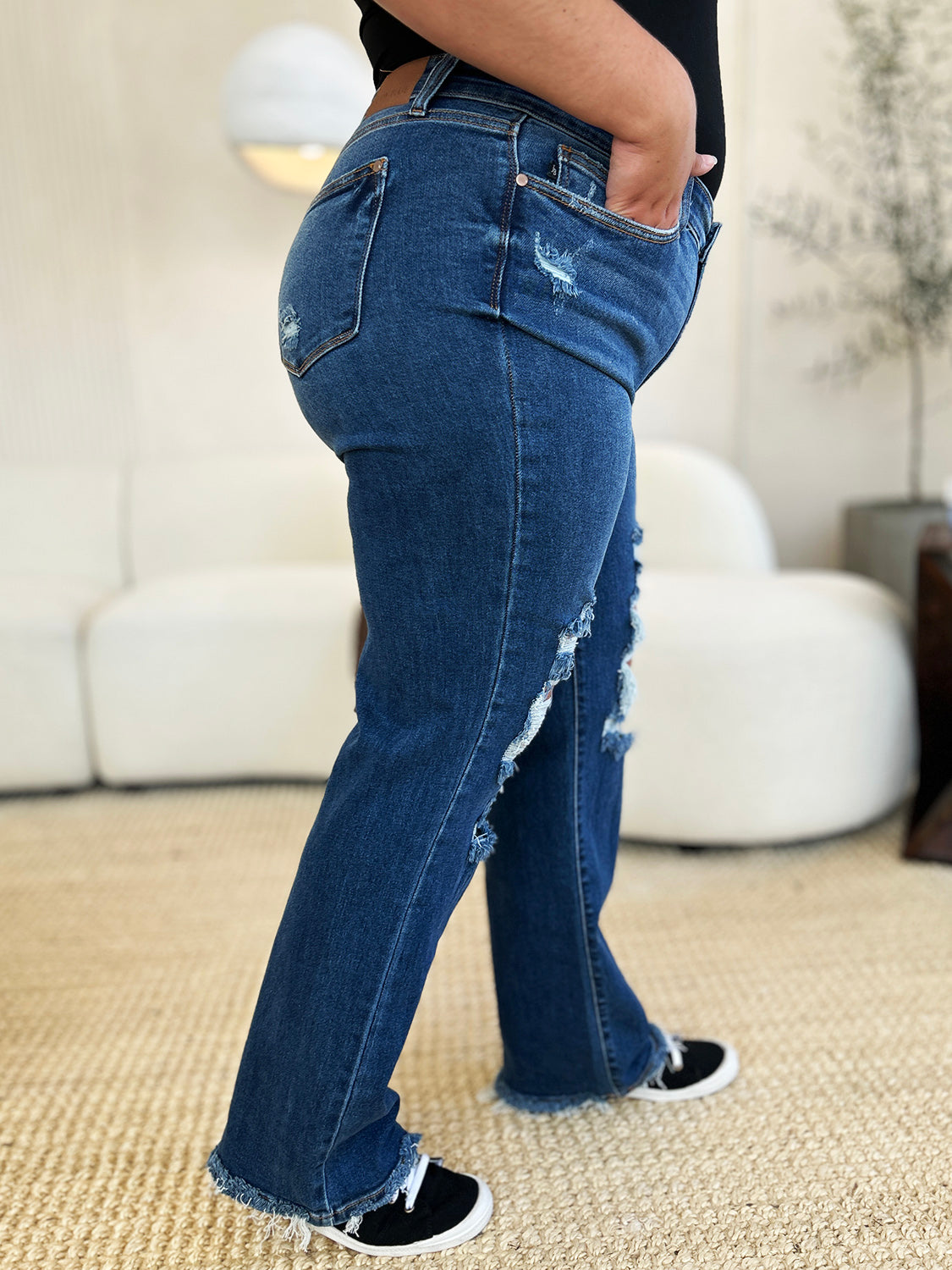 Person standing indoors wearing Judy Blue Full Size Mid Rise Distressed Raw Hem Jeans and black sneakers.