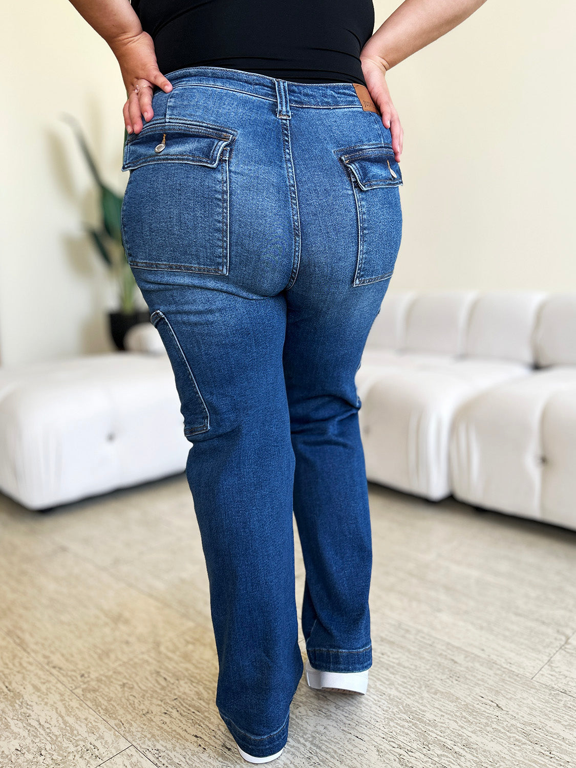 A person wearing Judy Blue Full Size High Waist Straight Cargo Jeans made of durable denim fabric and a white top walks on a wooden floor, with a white sofa and green plant in the background.