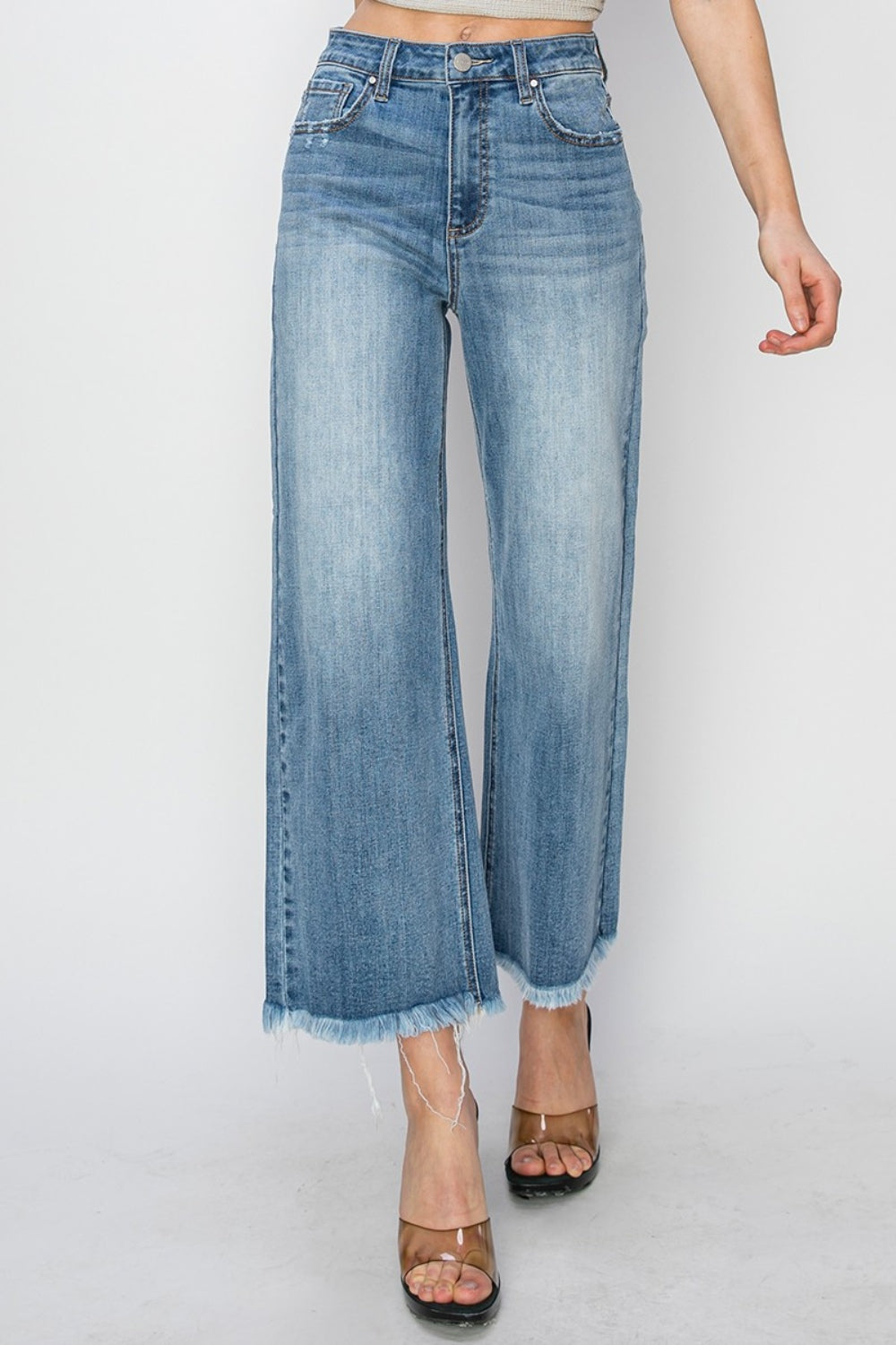 Person wearing RISEN Raw Hem Cropped Wide Leg Jeans in blue with a beige crop top. Only the lower half of the body is visible, paired with strappy black heels.