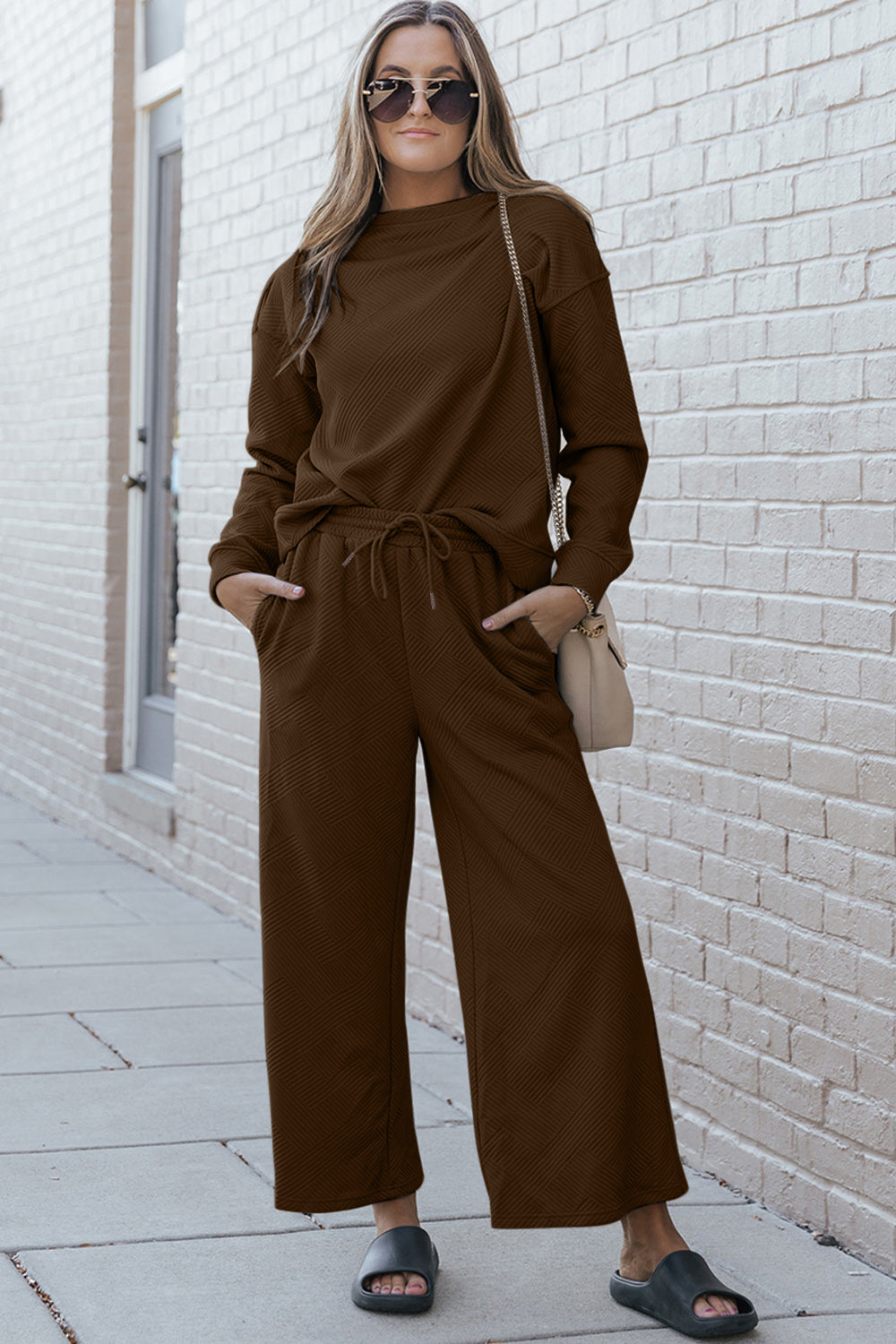 A woman with blonde hair, dressed in the Double Take Full Size Textured Long Sleeve Top and Drawstring Pants Set featuring a stretchy black long-sleeve top and matching wide-leg trousers made of rayon spandex, stands barefoot in a room with white walls and a wooden floor.