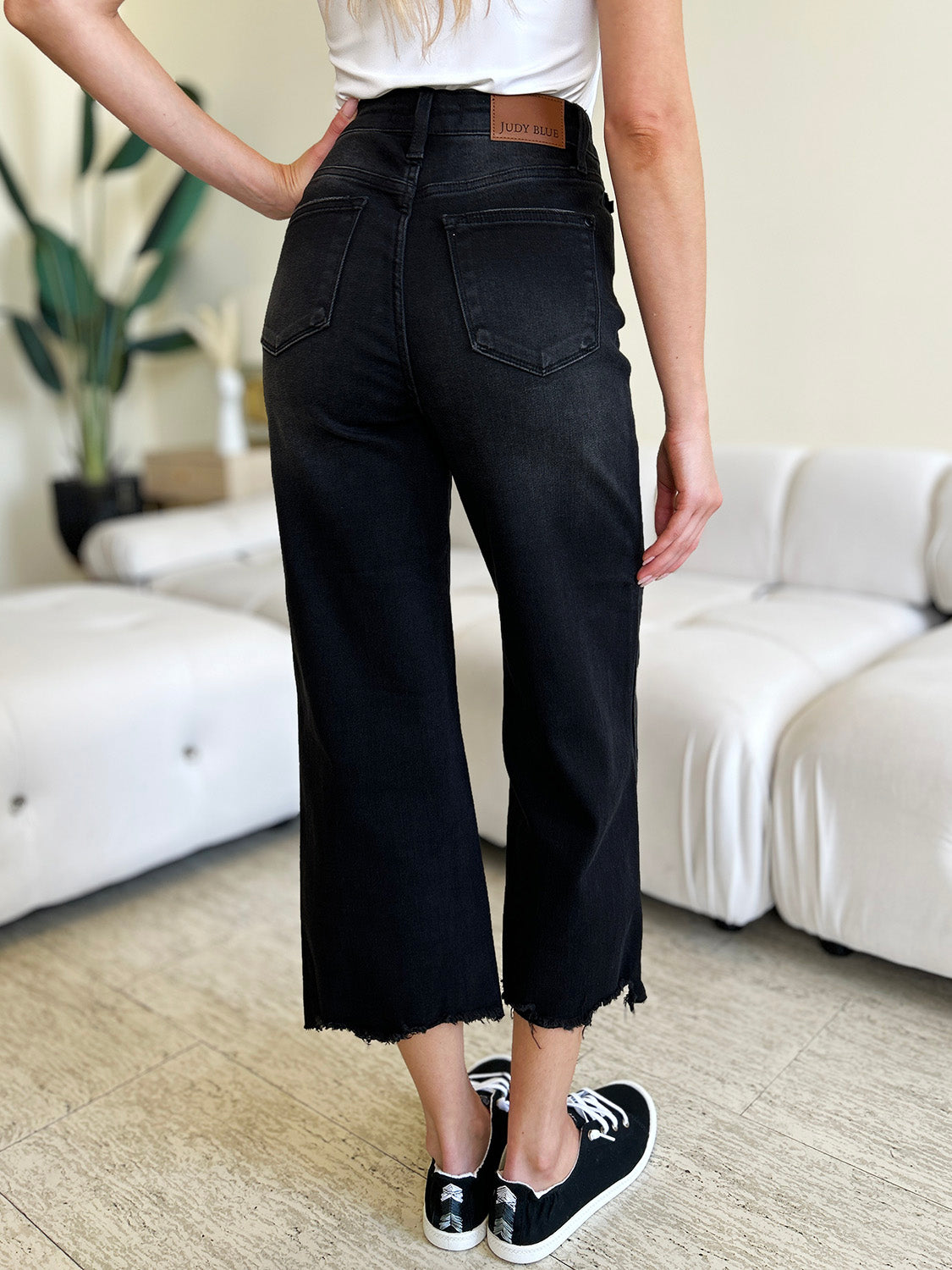 Person wearing Judy Blue Full Size High Waist Button Fly Jeans and black sneakers, standing indoors. Hands are in pockets, and the background includes a white couch and a plant. These high-waisted jeans make the outfit sleek and stylish.