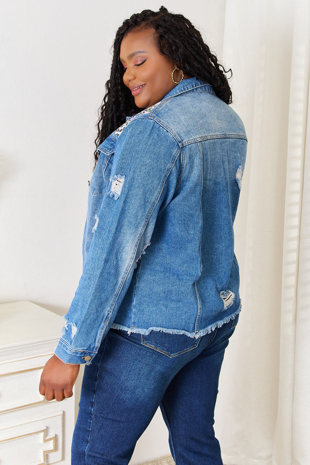 A person smiles while wearing the Baeful Leopard Floral Distressed Raw Hem Denim Jacket with embroidered patches, paired with a white shirt and blue jeans, as they stand indoors near a white piece of furniture.