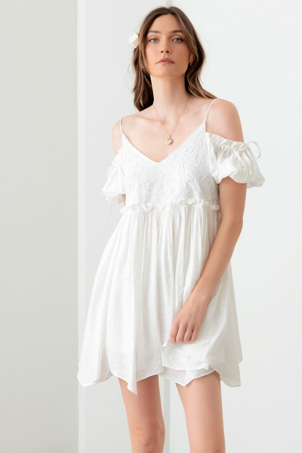 A woman poses in a boho-inspired, embroidered floral shirred cold shoulder mini dress by Mustard Seed. She accessorizes with a gold necklace and maintains a neutral expression against the backdrop of a plain white wall.