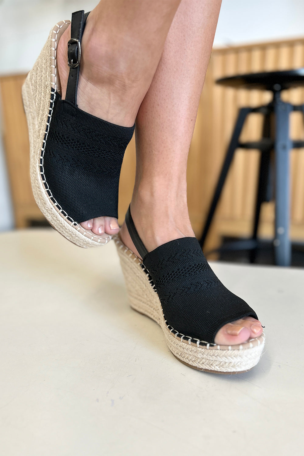 Close-up of a person's feet wearing chic black Forever Link Peep Toe High Heel Sandals with a braided espadrille base and adjustable heel straps. The background is blurred, making these sandals the stylish choice for any outfit.