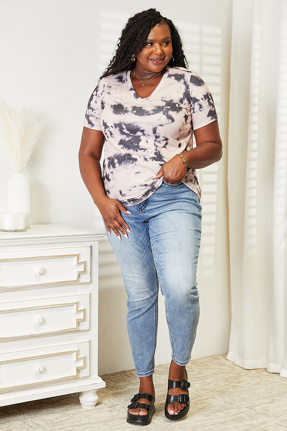 A person in a Double Take Tie-Dye V-Neck T-Shirt and jeans, epitomizing boho-chic, stands indoors, smiling at the camera. A vase with dried plants is visible in the background.