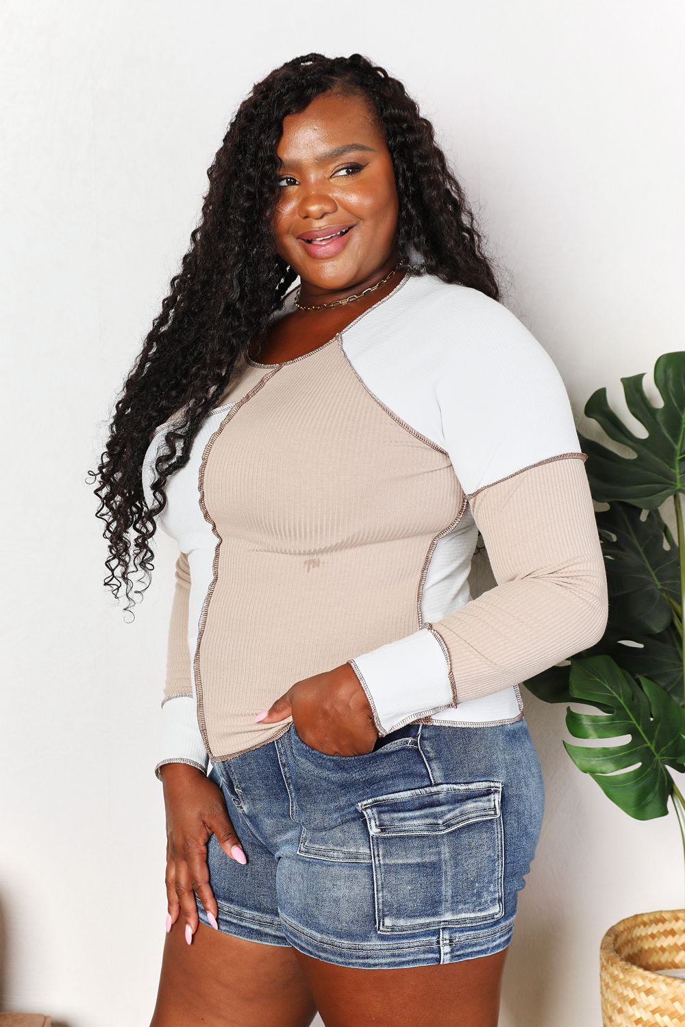 A woman with long brown hair is wearing the "Double Take Color Block Exposed Seam Top" in beige and white, paired with jeans. She stands with one hand in her pocket and the other touching her hair, smiling. A plant is visible in the background.