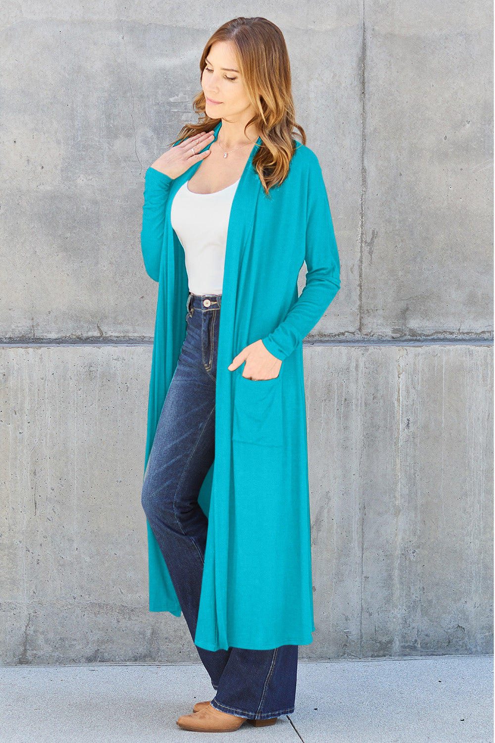 A woman stands against a concrete wall, wearing the Basic Bae Full Size Open Front Long Sleeve Cover Up made from rayon spandex, a white top, blue jeans in her perfect size, and brown shoes with her hands in her pockets. Machine wash cold for best results.