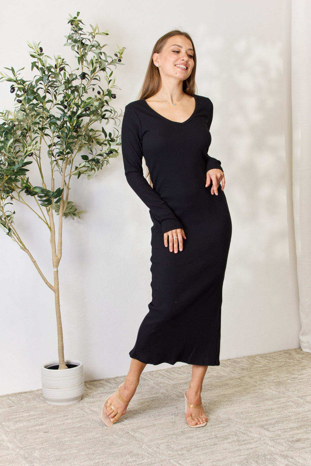 A person stands indoors wearing the chic Culture Code Full Size Ribbed Long Sleeve Midi Slit Dress paired with sandals, next to a potted plant.