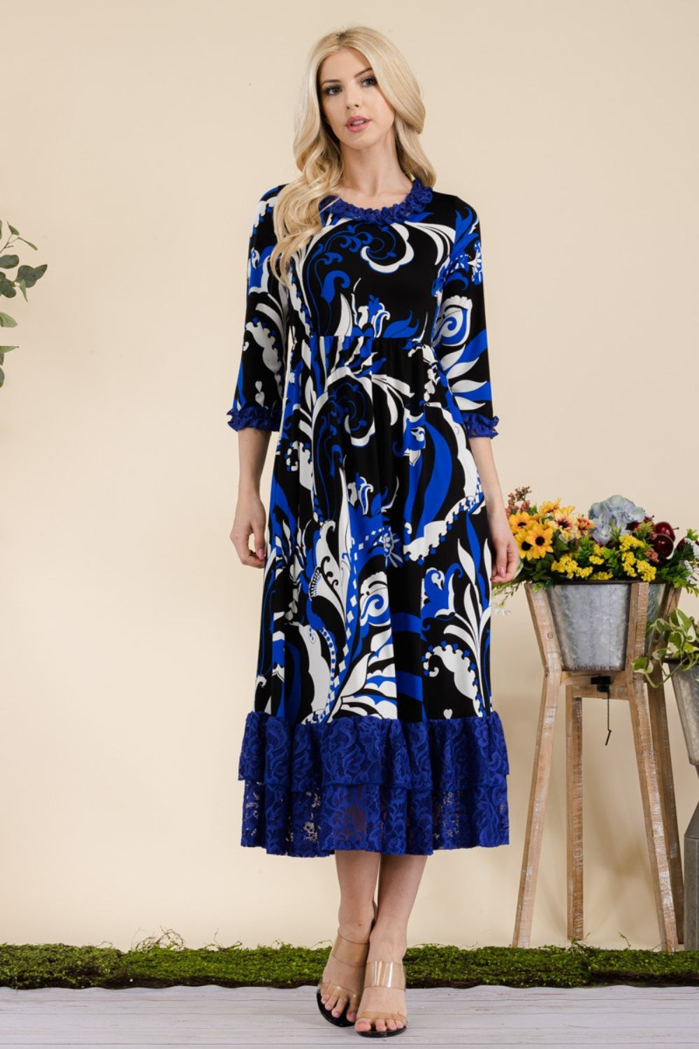 A woman wearing the Celeste Full Size Paisley Print Lace Ruffled Midi Dress stands near a wooden table adorned with a bucket of flowers.