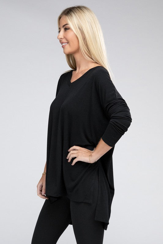 A woman with long, blonde hair is wearing a loose-fitting, brown Dolman Long Sleeve V-Neck Side Slit Hi-Low Hem Top and black pants. She poses with one hand on her hip and a neutral facial expression against a light grey background.