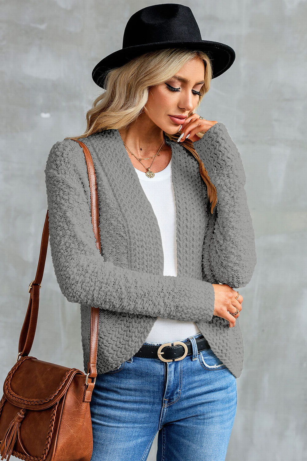 A woman in an Apricot Popcorn Knit Open Front Cardigan and jeans stands against a brick wall, looking to the side with a slight smile.