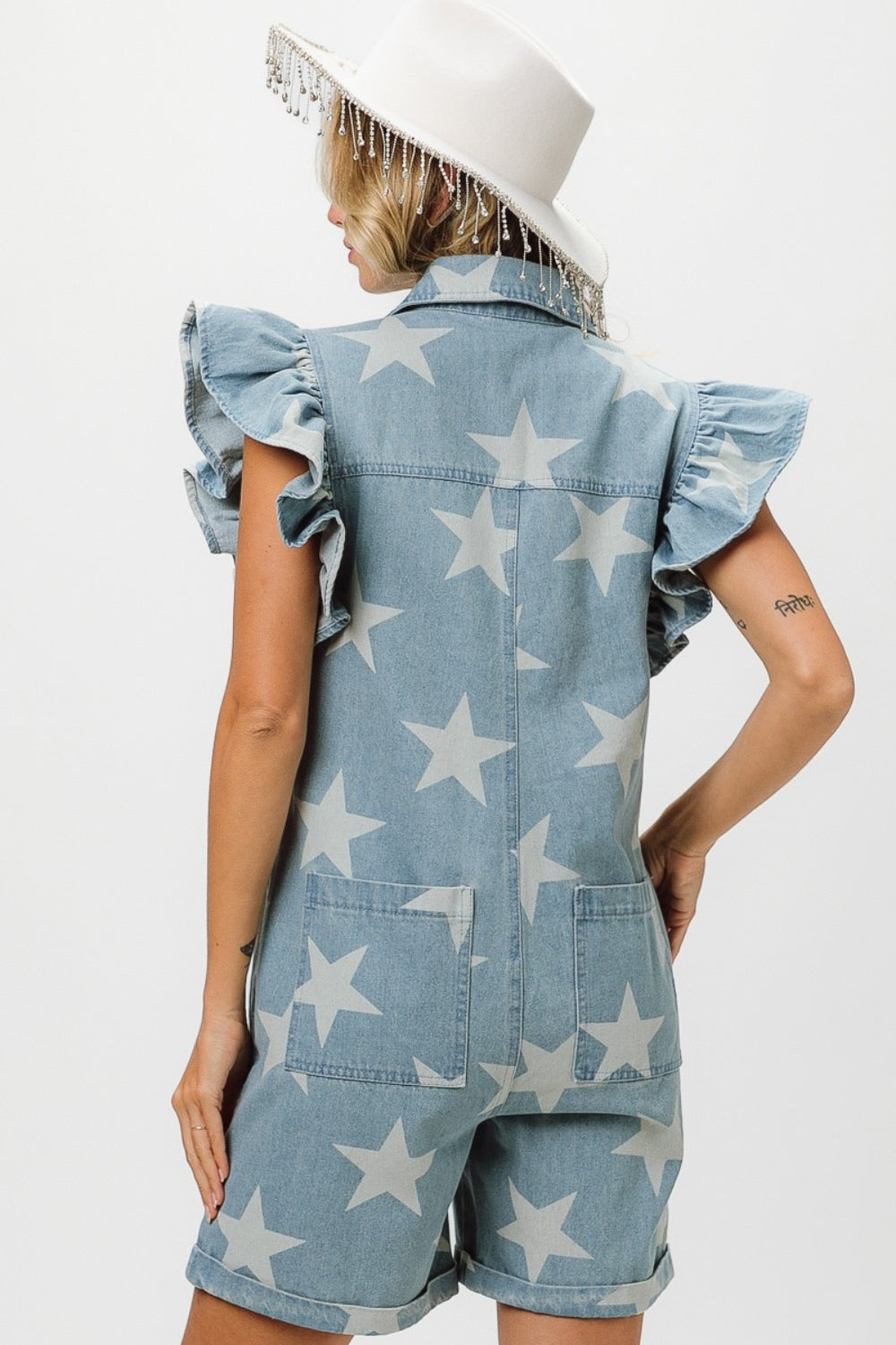 A person wearing the BiBi Ruffled Star Half Zip Denim Romper adjusts a beaded hat on their head.