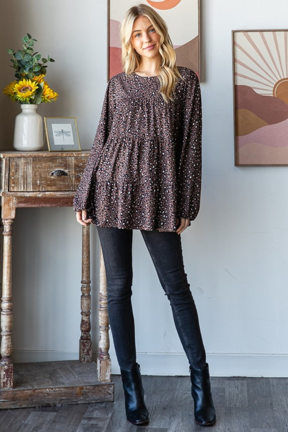 A woman wearing a Heimish Leopard Long Sleeve Tiered Blouse and black pants stands by a window indoors, looking at the camera—embodying a chic and feminine wardrobe.