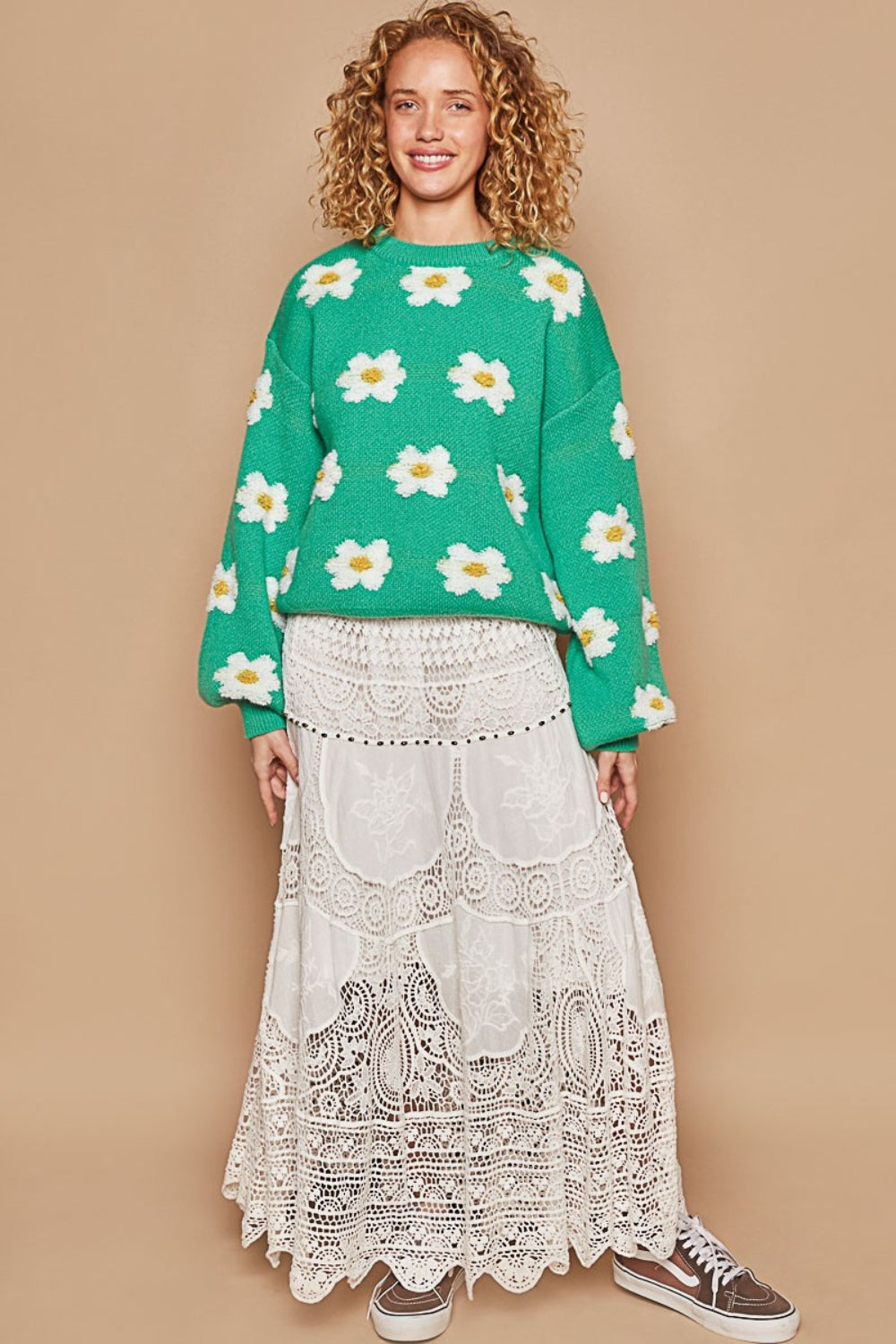 A person with curly hair wearing the POL Daisy Pattern Drop Shoulder Sweater, which is an oversized green sweater with a daisy pattern, and a lace skirt stands against a beige background, smiling.