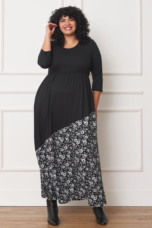 A woman with curly hair is wearing the Asymmetrical Leopard Accent Maxi Dress, featuring a long black design with a floral patterned section at the bottom. She stands against a white paneled wall, her hands tucked in her dress pockets, showcasing its elegant design.