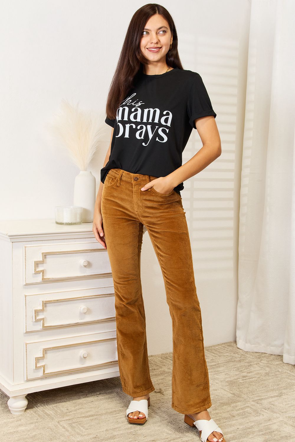 A woman wearing a Simply Love THIS MAMA PRAYS Graphic T-Shirt, paired with brown pants, stands gracefully in a softly lit room, exuding a sense of calm and spiritual gift.