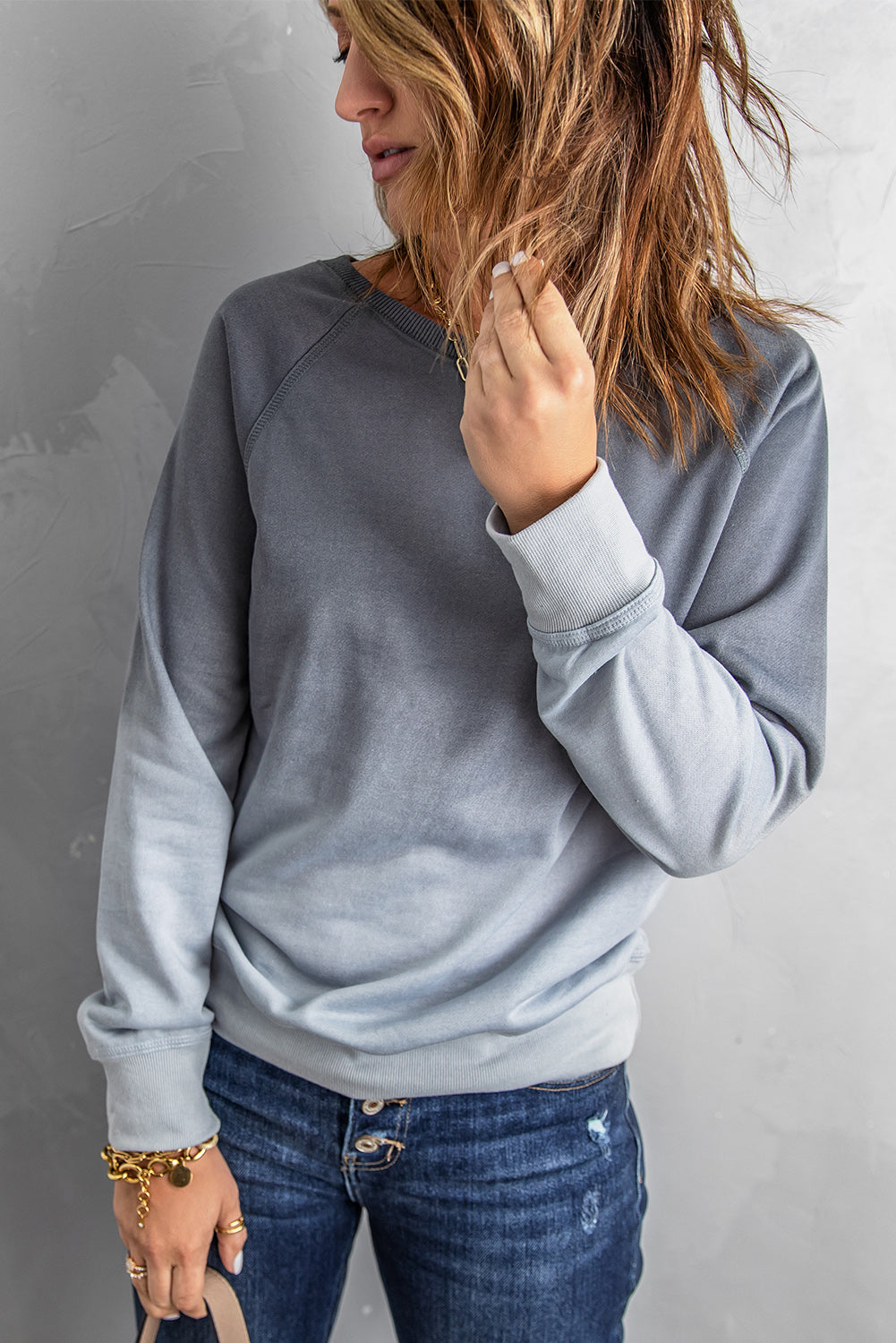 A person with wavy hair stands facing a gray wall, wearing a Gray Ombre Crewneck Long Sleeve Sweatshirt and blue jeans, with one hand on their hip.