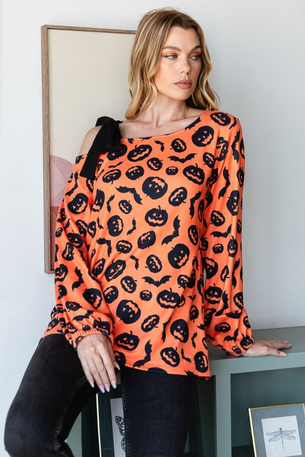 A woman smiles while modeling the Heimish Pumpkin Print One Shoulder T-Shirt, featuring an off-shoulder, long-sleeve design that is perfect for fall season fashion. She stands indoors with a plant and wall art in the background.