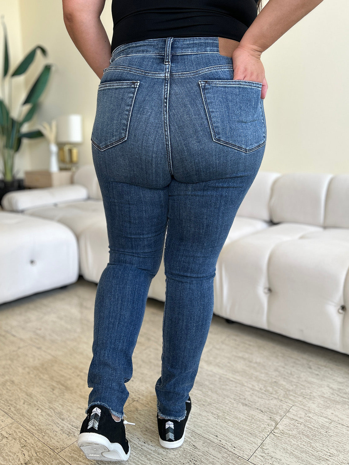 A person wearing Judy Blue Full Size High Waist Distressed Skinny Jeans and black sneakers stands indoors on a wooden floor, with a white sofa and green plant in the background.