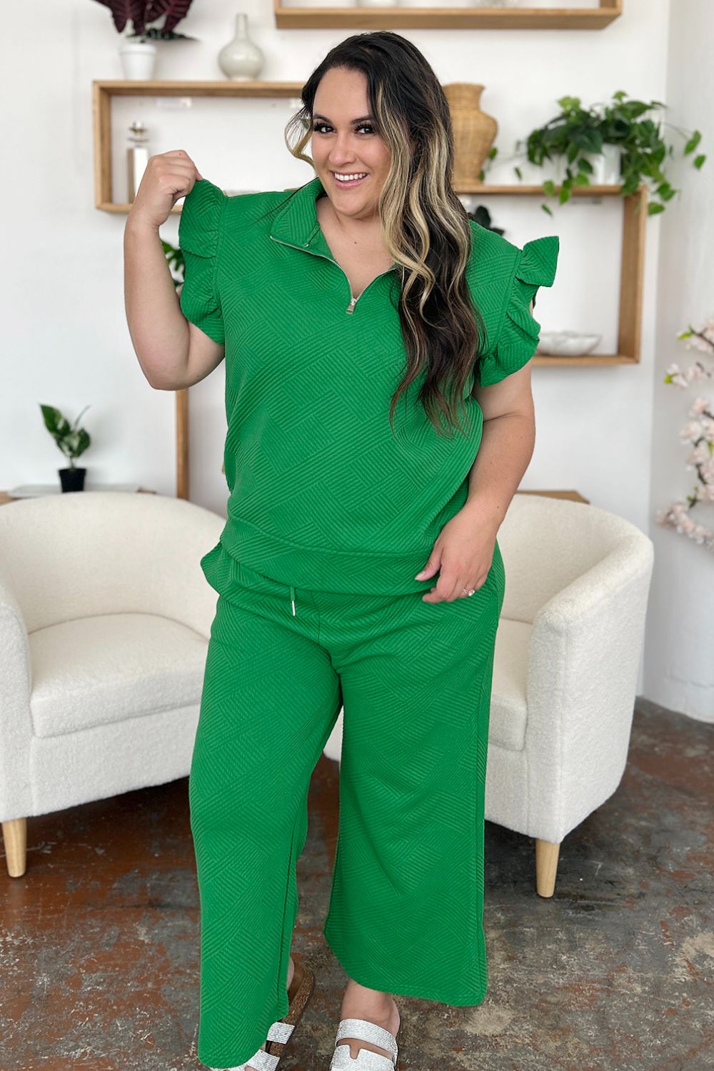 A woman stands wearing the Double Take Texture Ruffle Short Sleeve Top and Drawstring Wide Leg Pants Set in a slightly stretchy, light gray fabric with zipper detail. The two-piece outfit is complemented by black sandals, and she has one hand in her pocket. Machine wash cold for easy care.