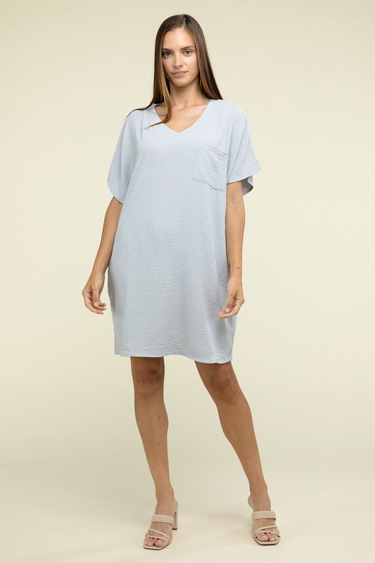 A woman wearing a loose light brown Woven Airflow V Neck T-Shirt Dress with Pockets stands against a plain background. Made from breathable fabric, this summer dress includes pockets where her hands rest as she smiles slightly.