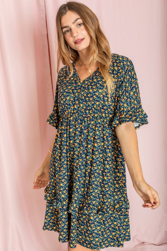 A woman wearing a Floral V Neck Ruffle Dress holds a wicker handbag, standing in a room decorated with a plant and partially visible furniture in the background.