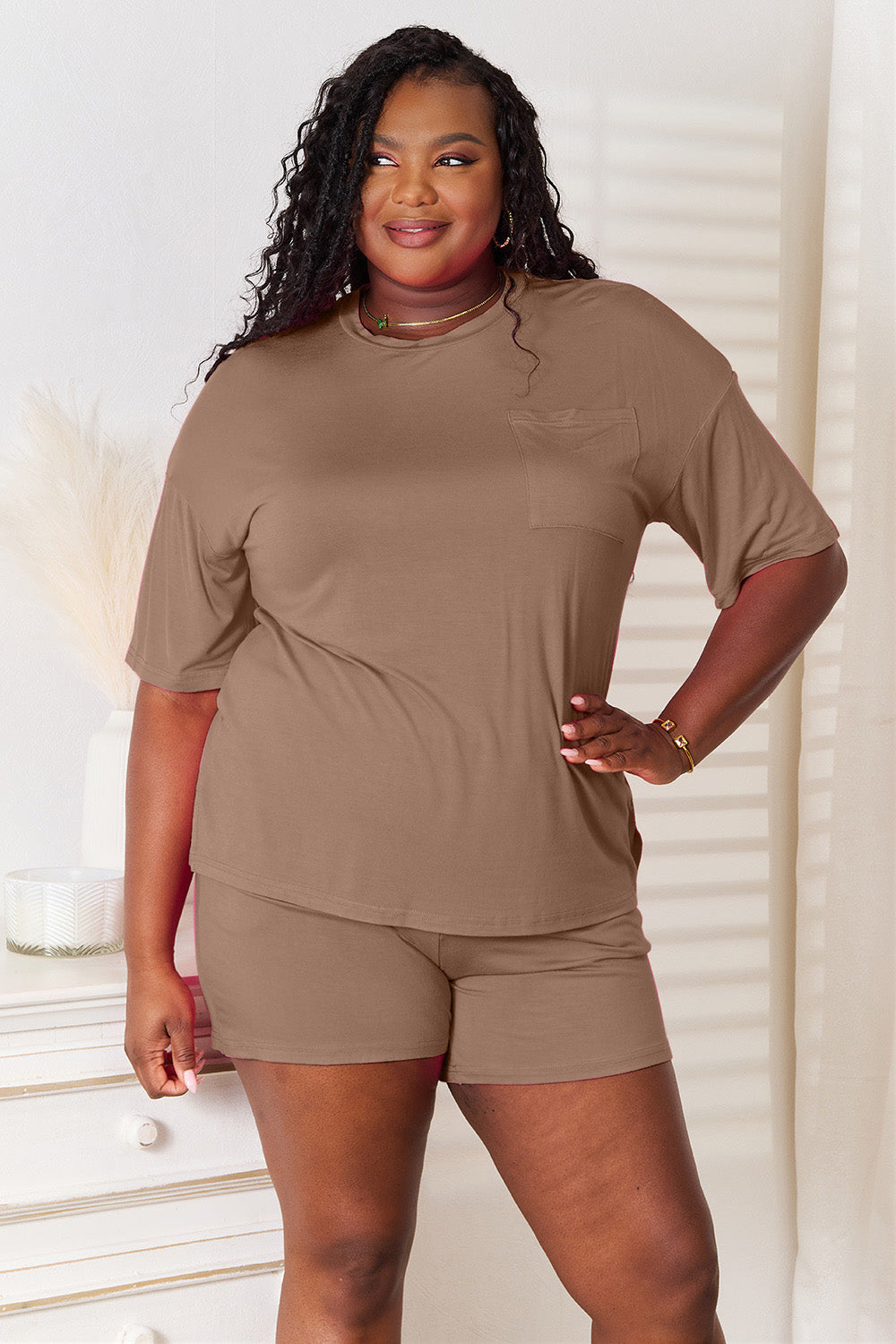 A woman is standing in a brightly lit room, wearing the Basic Bae Full Size Soft Rayon Half Sleeve Top and Shorts Set in a relaxed, stretchy turquoise fabric. She has one hand placed in her pocket and is smiling while looking slightly to the side.