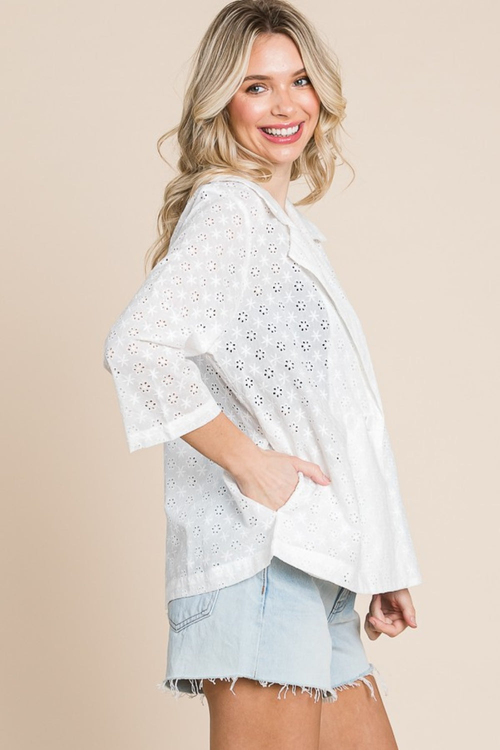 A woman stands against a beige background, wearing the chic Culture Code Double Breasted Eyelet Jacket with Pockets over a black top and light denim shorts.