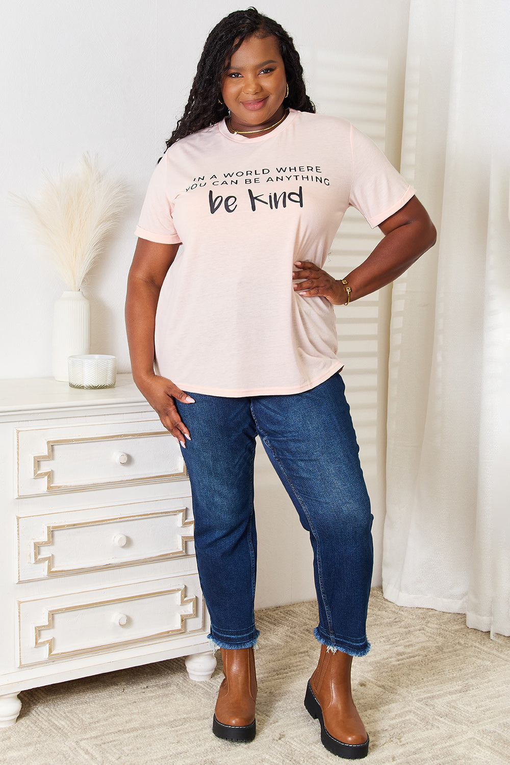 A person wears a Simply Love Slogan Graphic Cuffed T-Shirt in light pink, adorned with the inspiring message, "In a world where you can be anything, be kind," and featuring stylish cuffed sleeves.