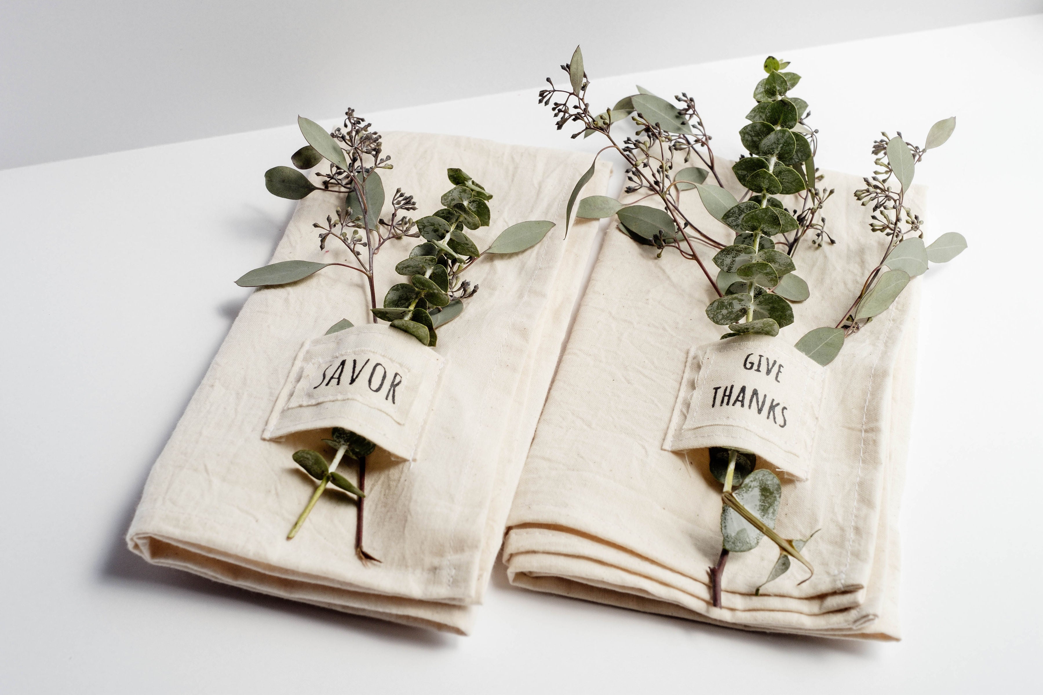 Pocket napkin set of four, skillfully crafted from up-cycled fabric, folded with a eucalyptus sprig and wooden ring, labeled "SAVOR.