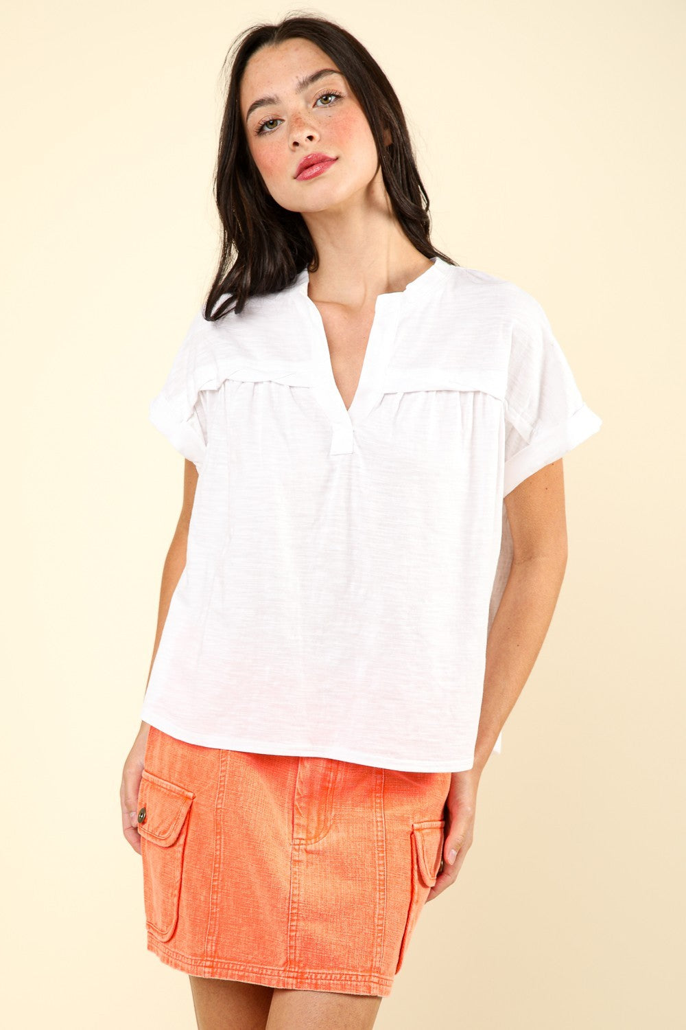 A woman wearing a white, VERY J Notched Short Sleeve Washed T-Shirt paired with an orange skirt poses against a neutral background, showcasing a versatile wardrobe staple.