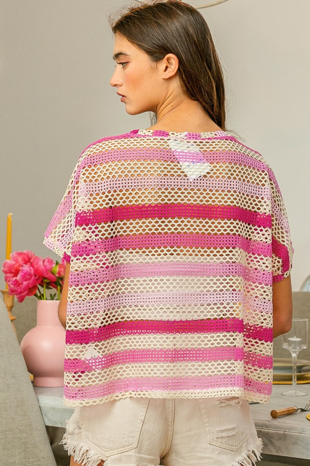 A woman wearing the BiBi Striped Openwork Short Sleeve Knit Cover Up in pink and white stands with one hand in her hair, surrounded by modern decor.