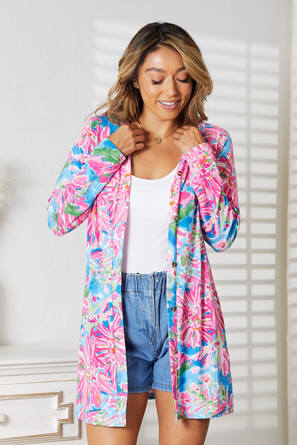 Wearing the Double Take Floral Open Front Long Sleeve Cardigan in shades of pink and blue over a white top and denim shorts, a person exudes feminine charm while standing in a well-lit room, smiling.