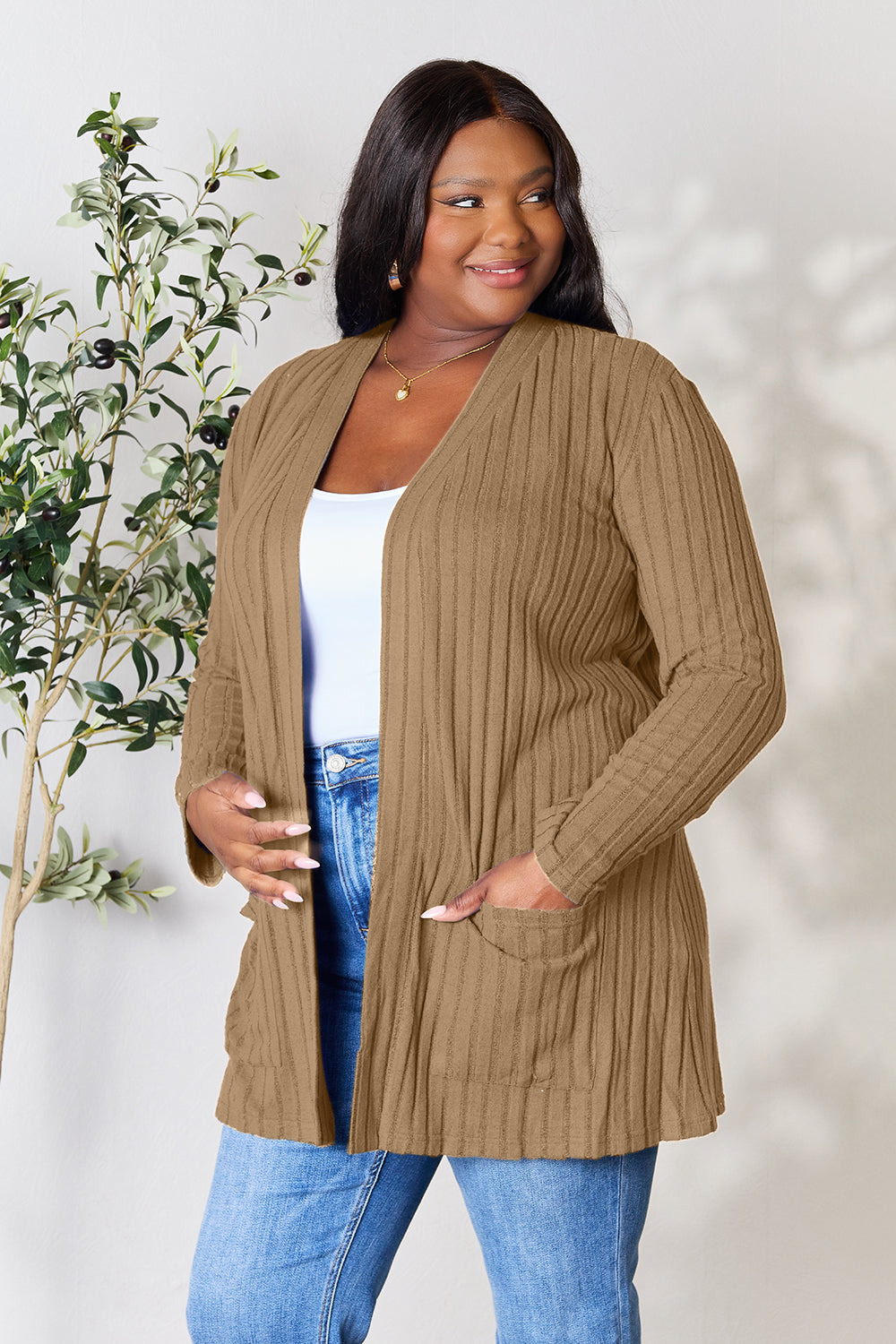 A woman in a Basic Bae Full Size Ribbed Open Front Cardigan with Pockets and jeans, made from stretchy material, stands in front of a plant, smiling slightly and looking to the side.