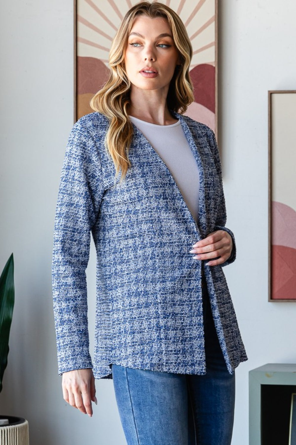 A woman with wavy blonde hair wears the Heimish Full Size Houndstooth Tweed Open Front Blazer in blue, paired with a white top and blue jeans. She stands in front of a wall adorned with framed art.